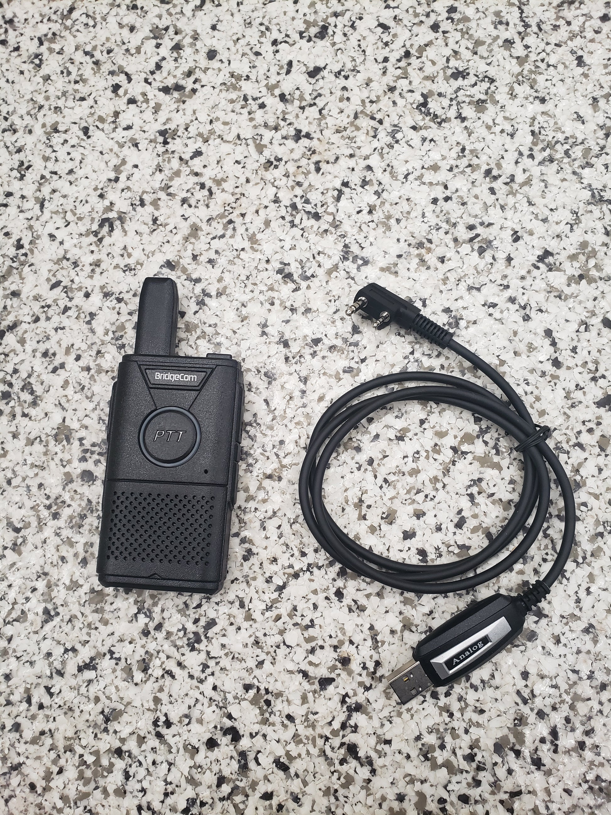 A black Buddy FRS handheld radio and the BridgeCom Buddy Programming Cable rest on a speckled gray countertop. The radio displays a "PTT" button, while the cable features a USB connector and right-angle plug for easy programming.