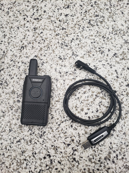 A black Buddy FRS handheld radio and the BridgeCom Buddy Programming Cable rest on a speckled gray countertop. The radio displays a "PTT" button, while the cable features a USB connector and right-angle plug for easy programming.