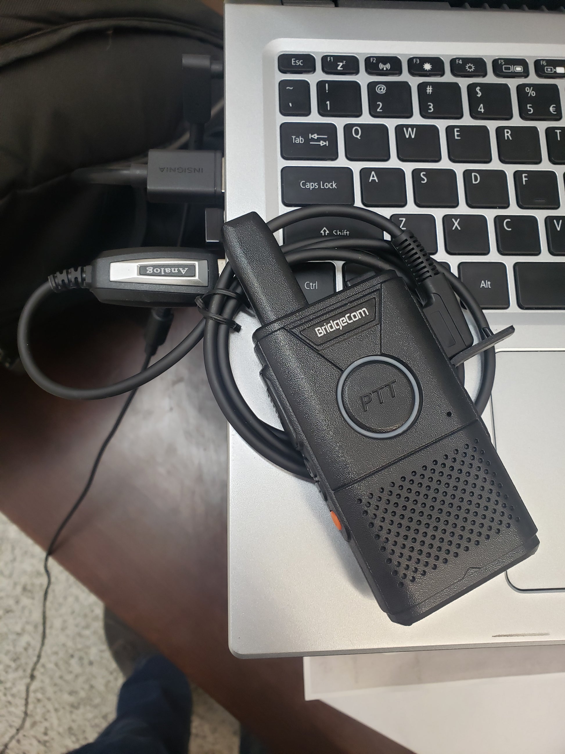 A BridgeCom Buddy Programming Cable with a coiled wire and earpiece is placed on the corner of an open laptop keyboard; part of a backpack and a laptop charger appear in the background.